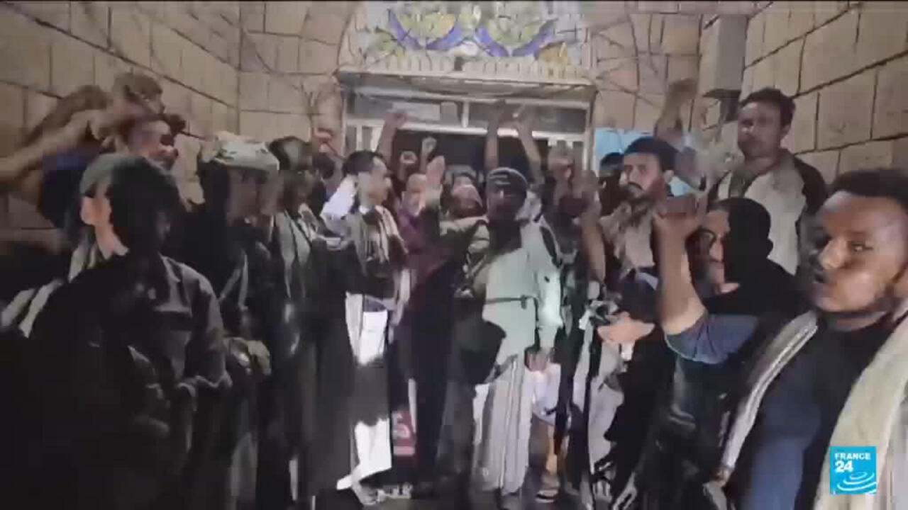Crowd gathered indoors, raising hands near a building entrance as others look toward the camera.