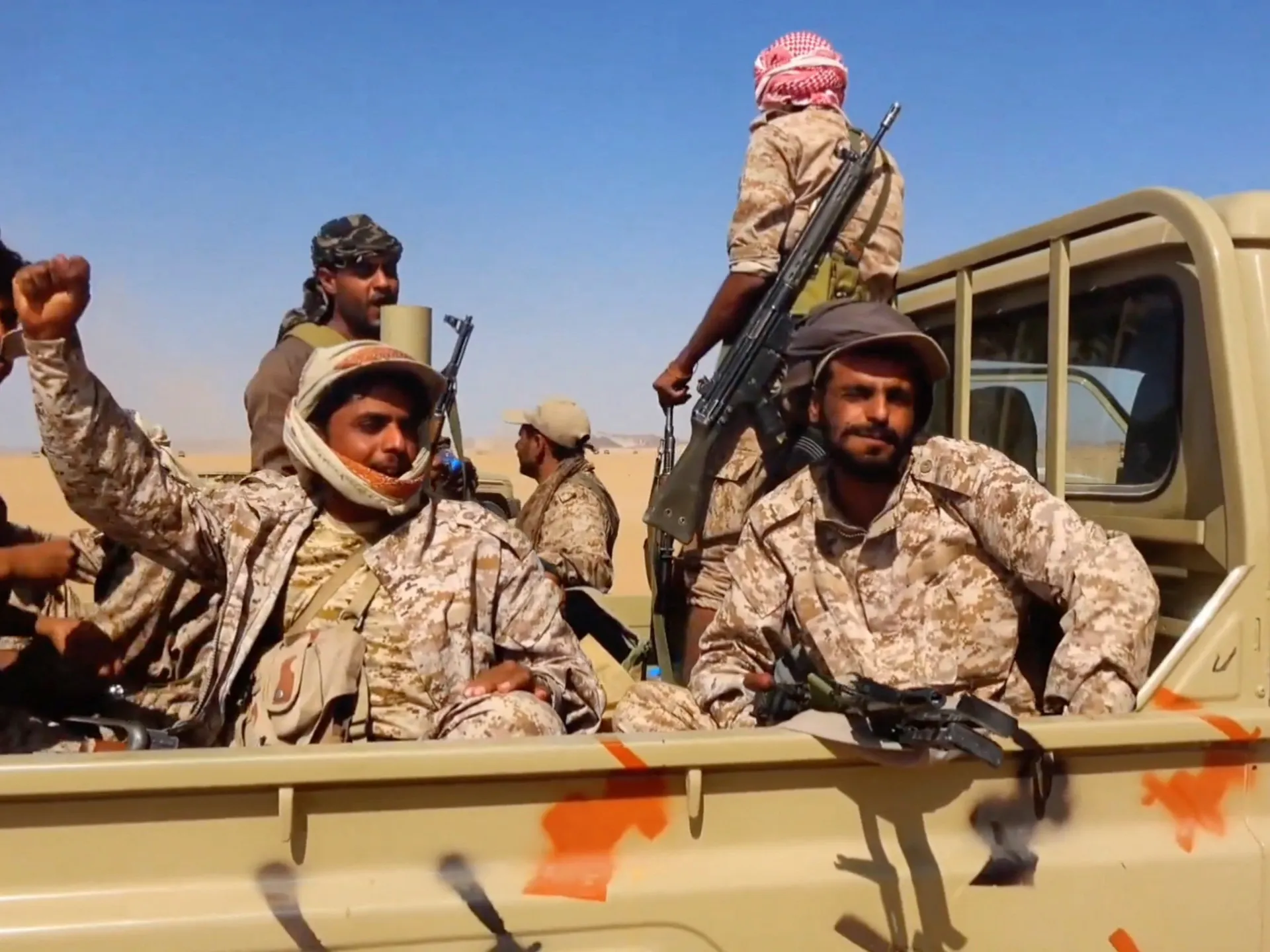 Armed men in desert camouflage ride in the back of a pickup truck in a desert.