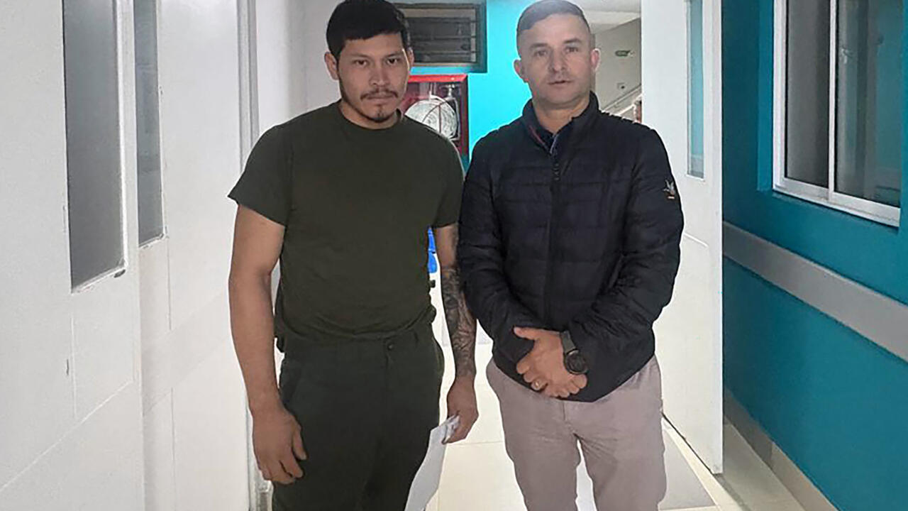 Two men stand in a brightly lit indoor corridor, facing the camera.