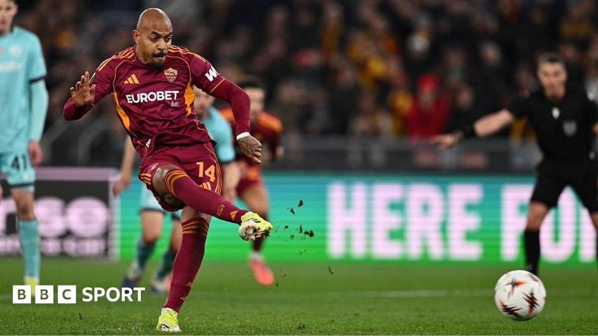 Footballer in burgundy kit strikes the ball during a match as opponents watch nearby
