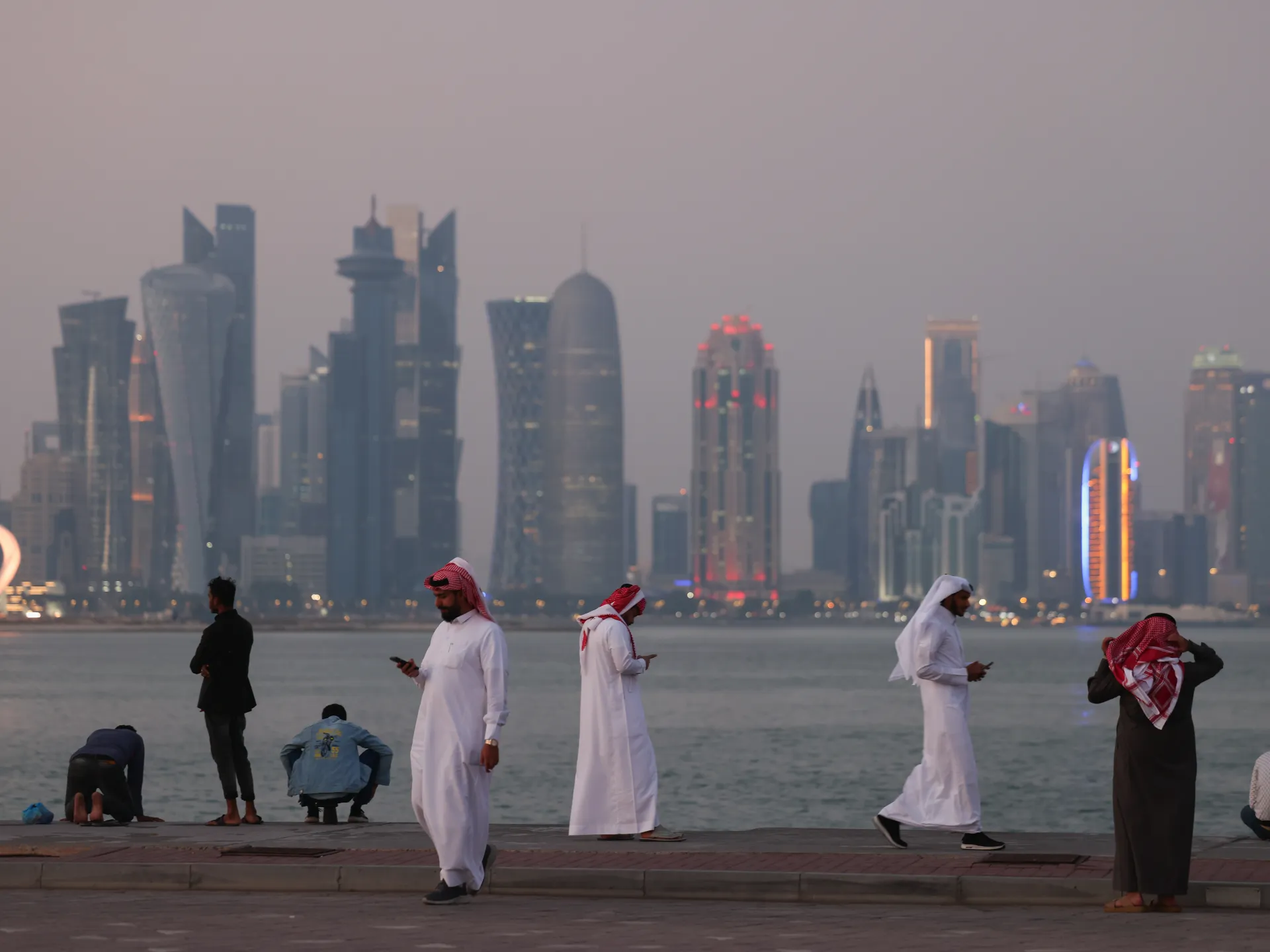 People gather along a waterfront promenade with a hazy illuminated skyline in the background.