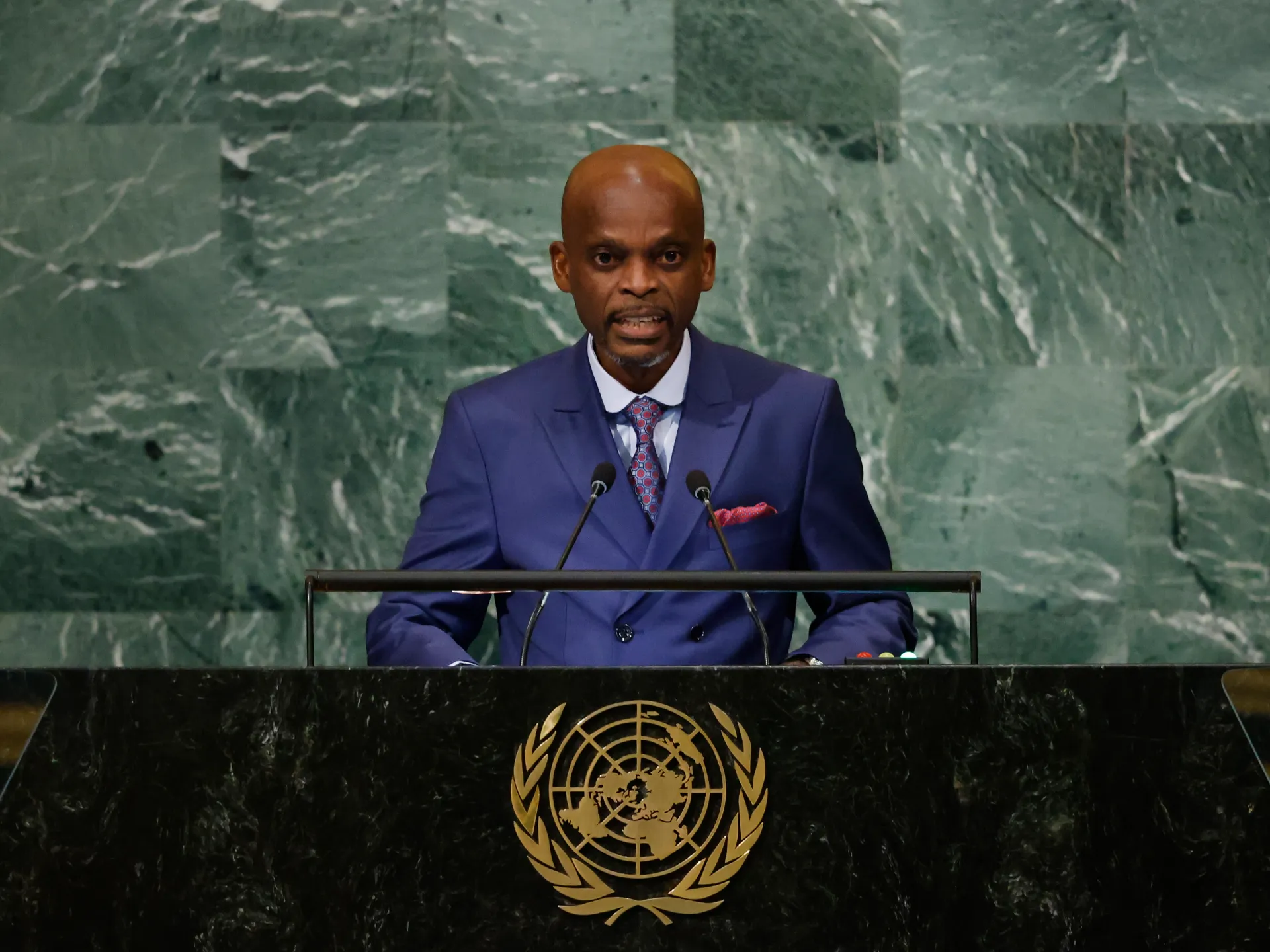 Man in blue suit speaking at a podium before a green marble wall