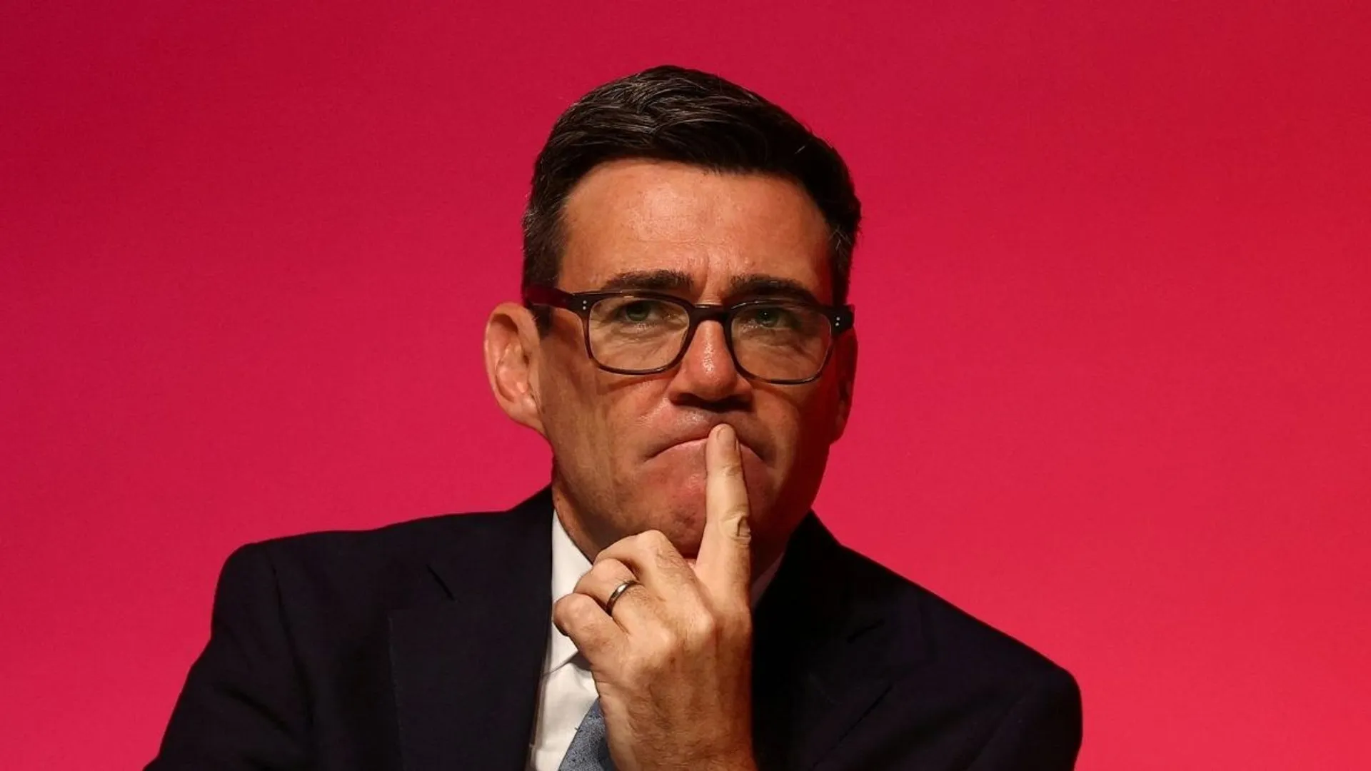 Man in glasses and dark suit touches his lips against a bright pink background.