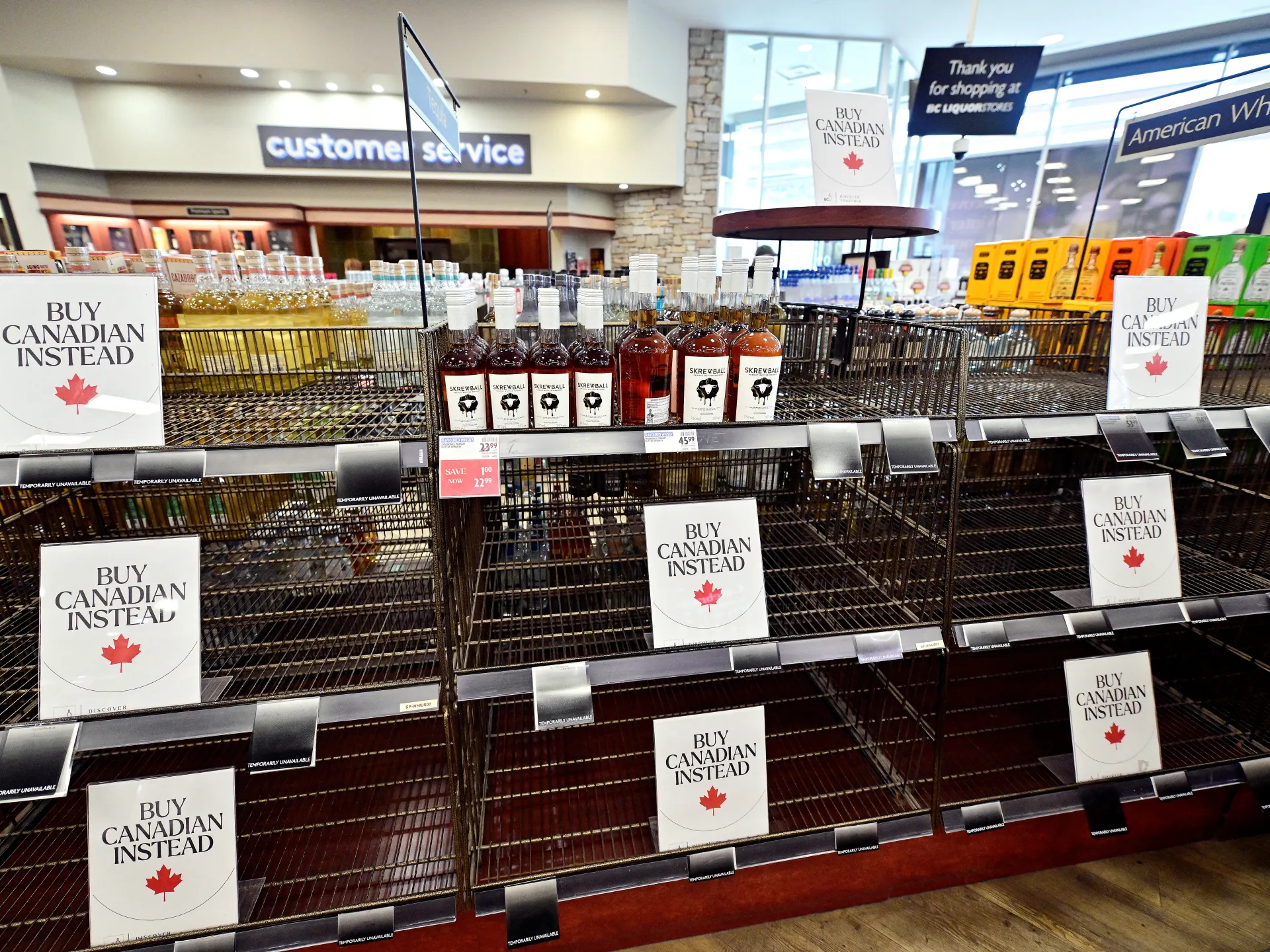 Liquor store shelves display Canadian-buying signs beside largely empty alcohol racks.