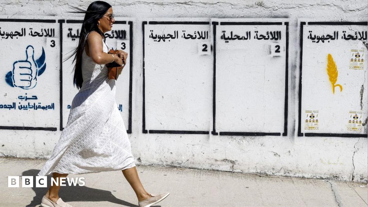 Woman in white dress walking past Arabic election posters painted on a weathered wall.