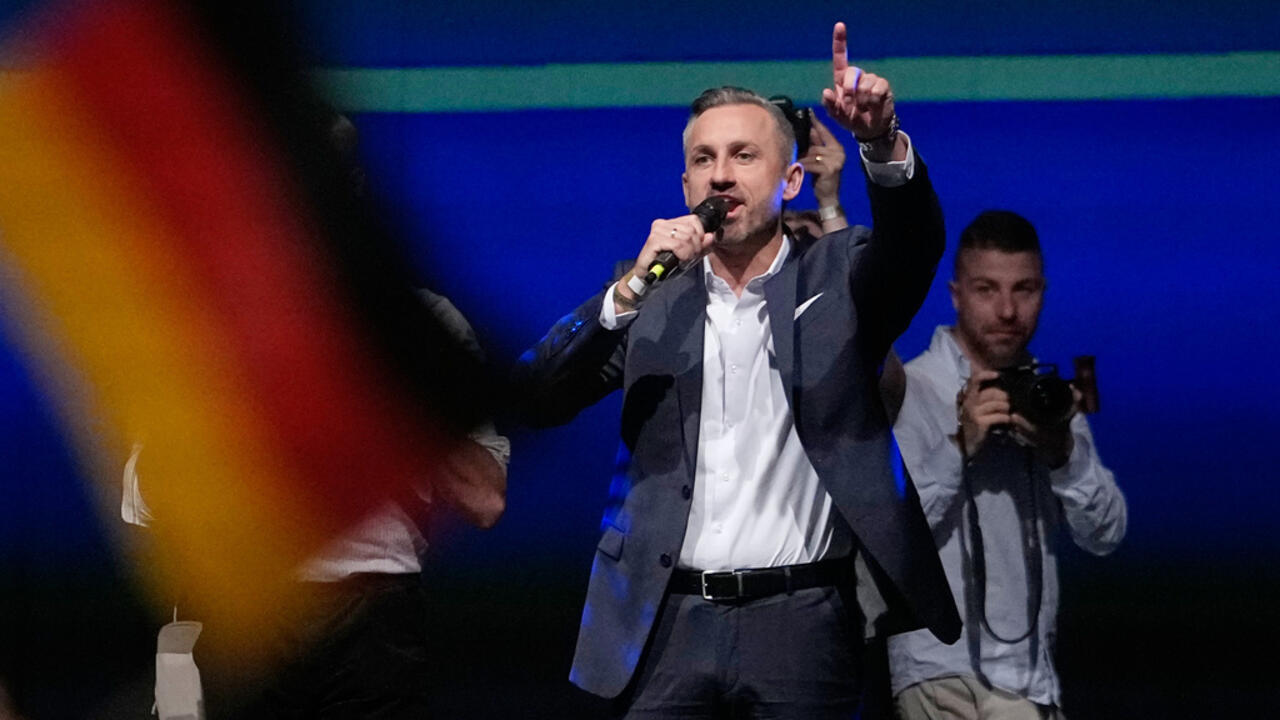Man in suit speaks into a microphone and raises one finger on a blue-lit stage.