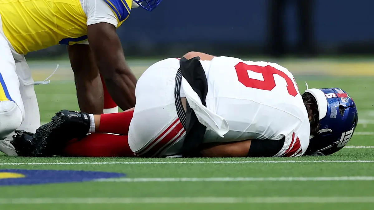 Injured football player in white uniform lies on field as another player kneels nearby.