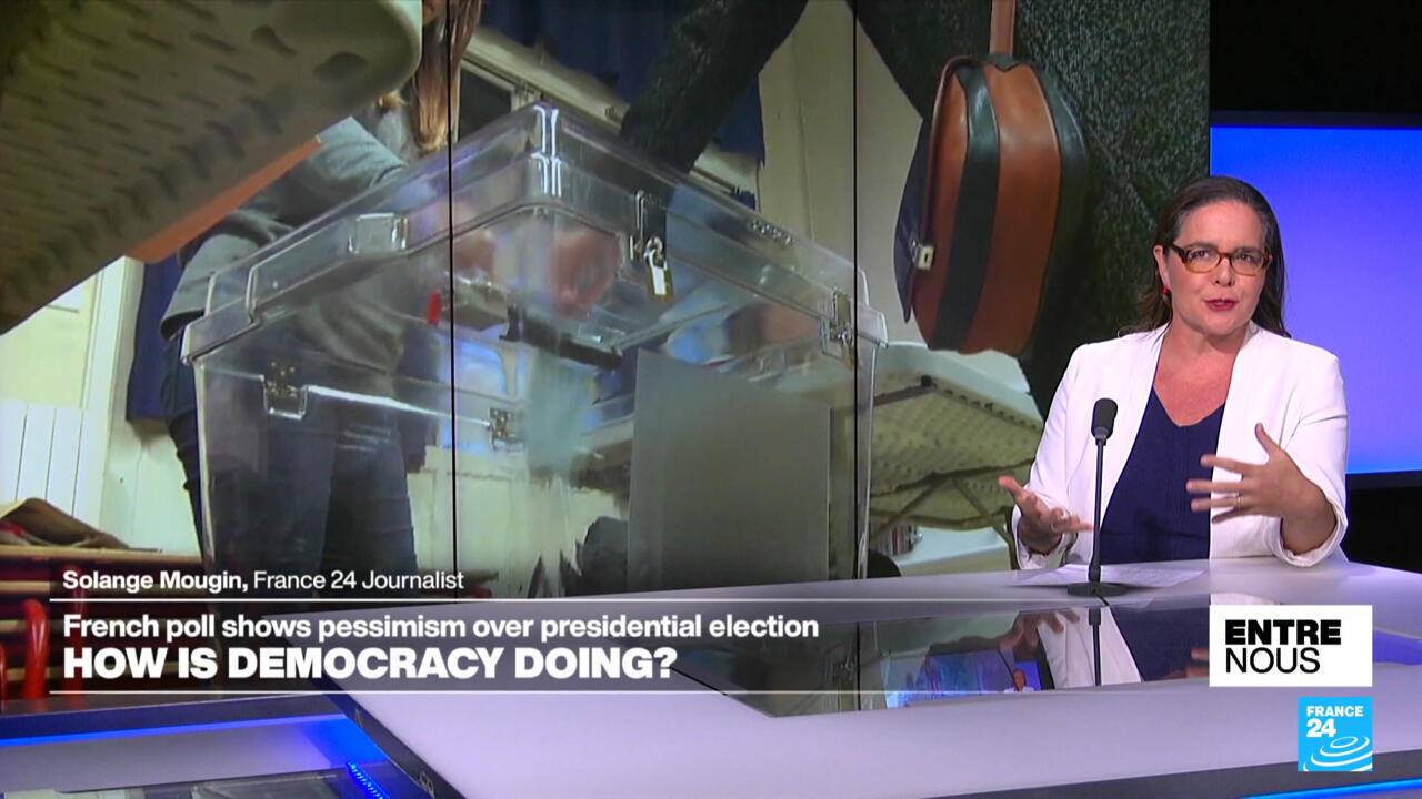 Woman speaks at a desk beside a ballot box, with election imagery displayed behind her