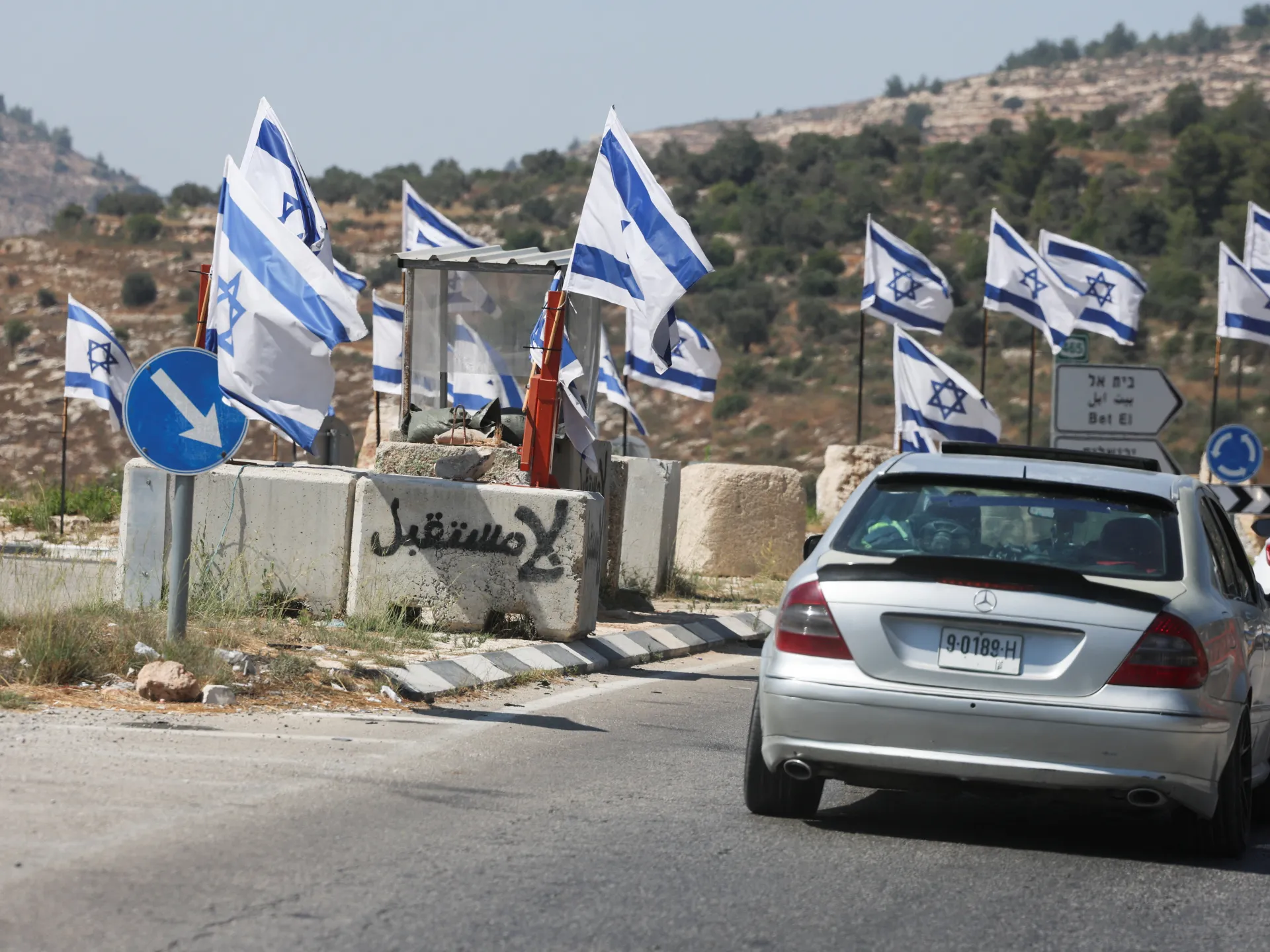 Israeli flags line a road beside concrete barriers, directional signs, and a silver Mercedes.
