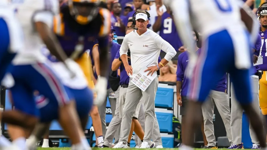 Football coach in white LSU attire stands on sideline surrounded by players during a game.