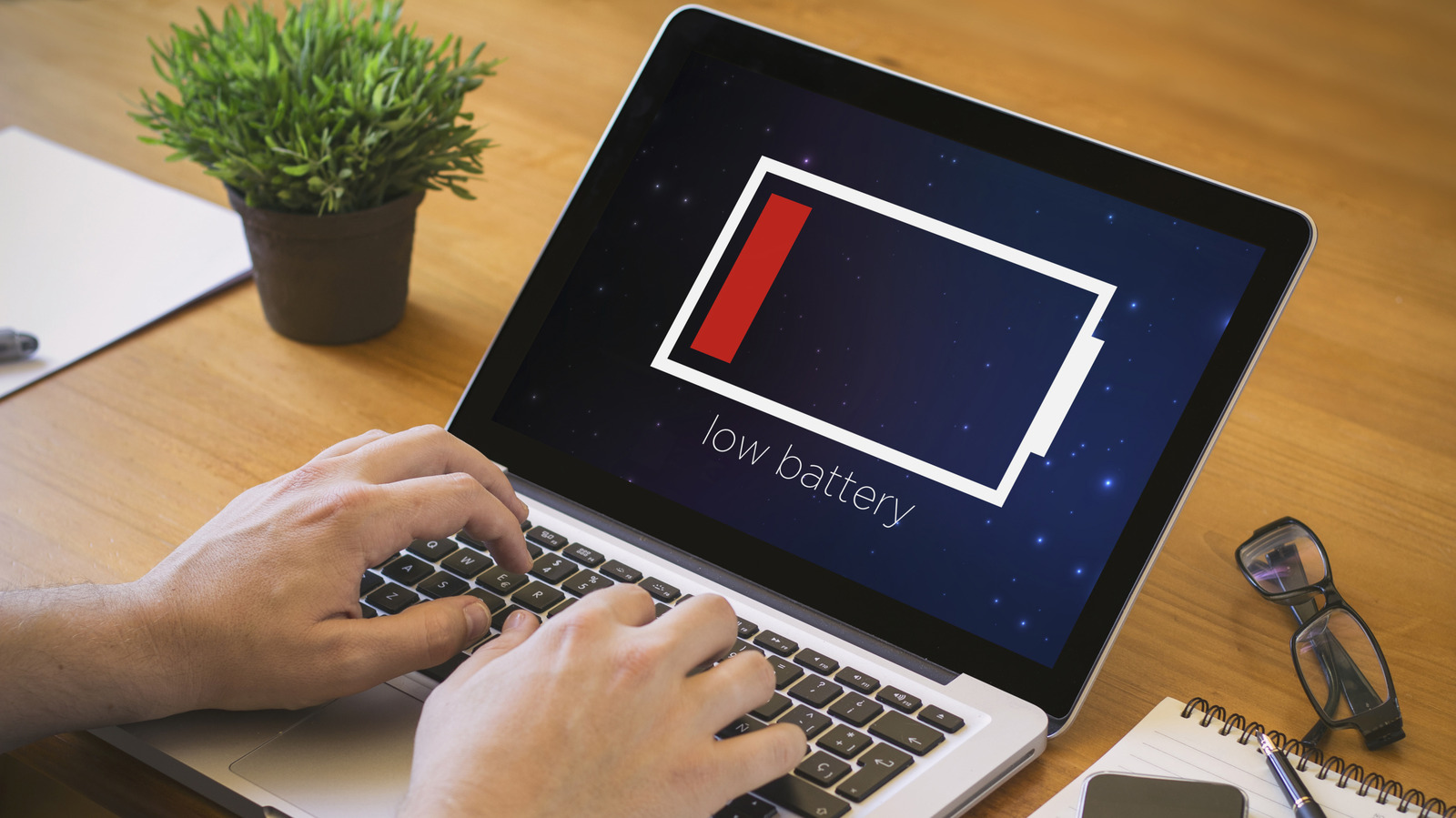 Person typing on a laptop displaying a low battery warning beside a potted plant and glasses