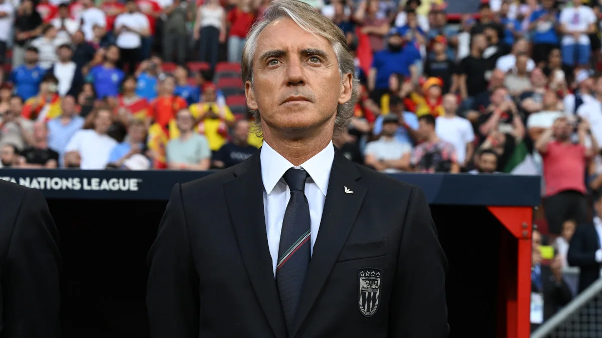 A gray-haired man in a dark suit stands before a crowded football stadium, looking ahead.