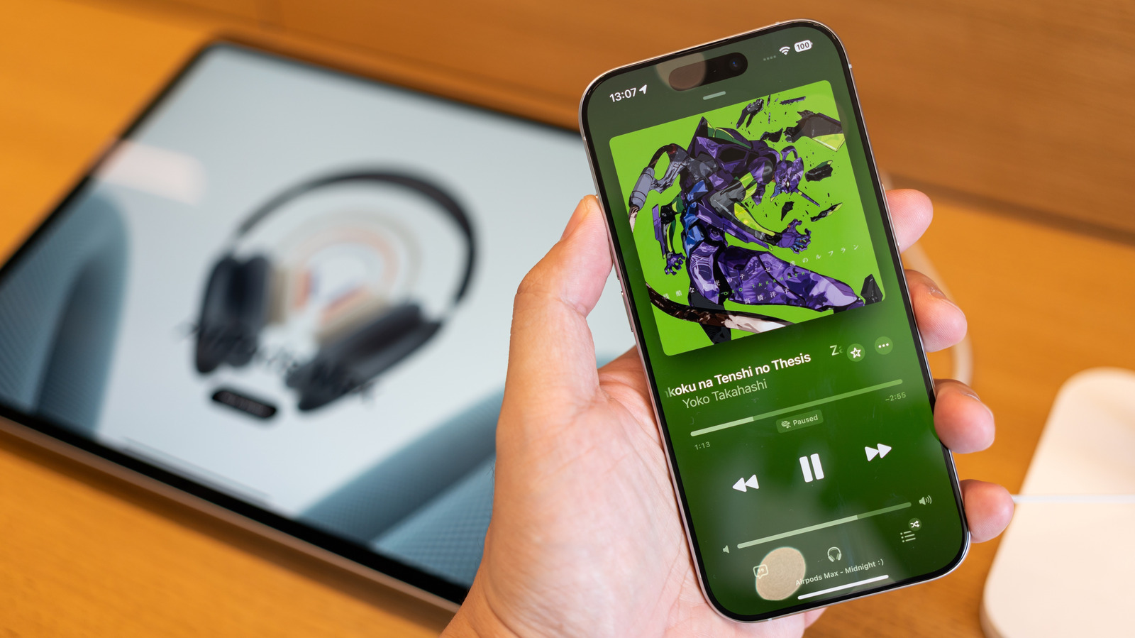 A hand holds a smartphone displaying Apple Music beside a blurred tablet and headphones.