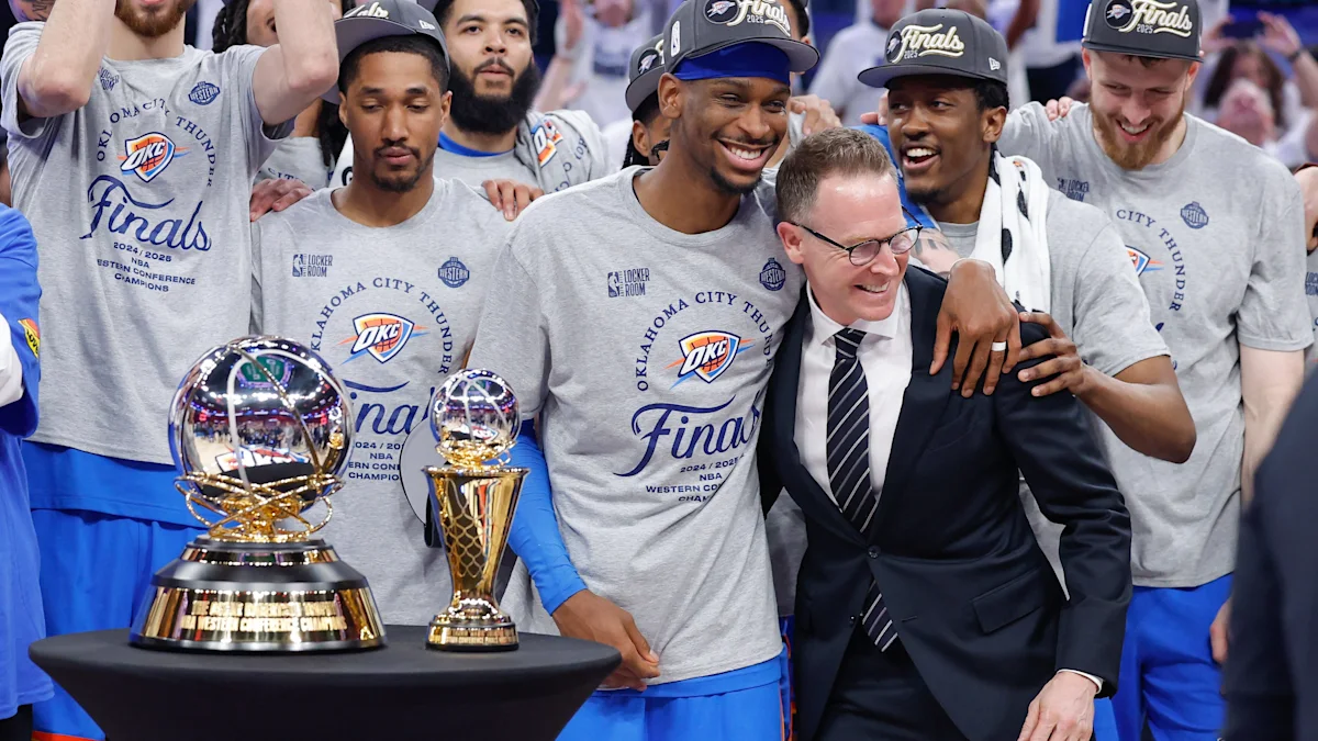 Basketball players in championship shirts pose with trophies as a suited man smiles among them.