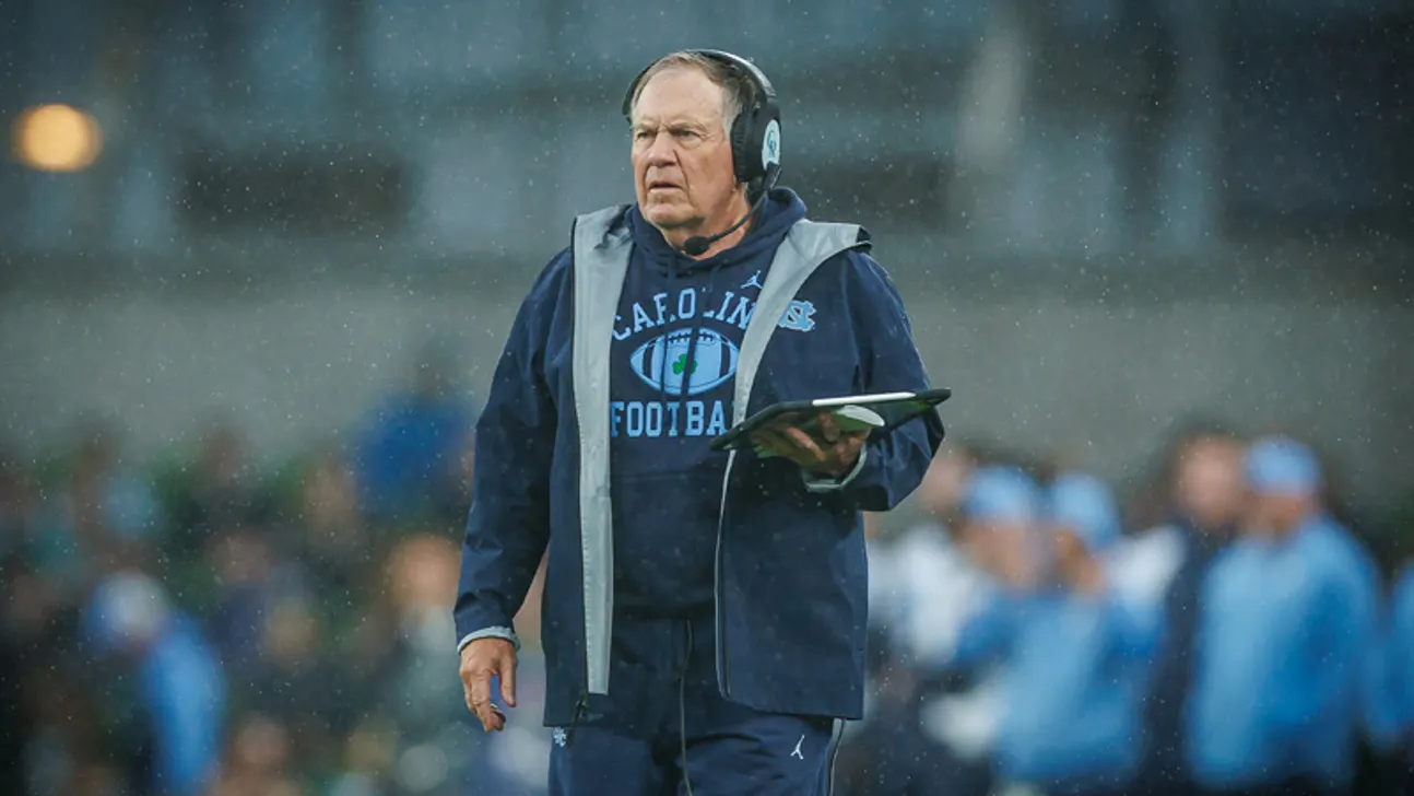 Football coach in headset and rain gear holds a tablet during a rainy game.