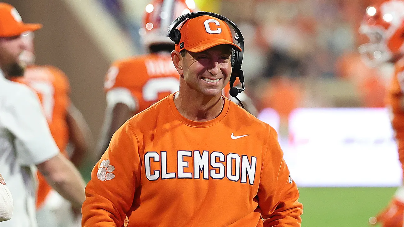Clemson coach in orange sweatshirt and cap wearing a headset on the football sideline