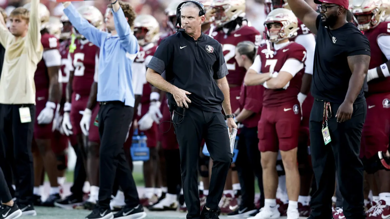 Football coach wearing headset stands on sideline among maroon-uniformed players and staff