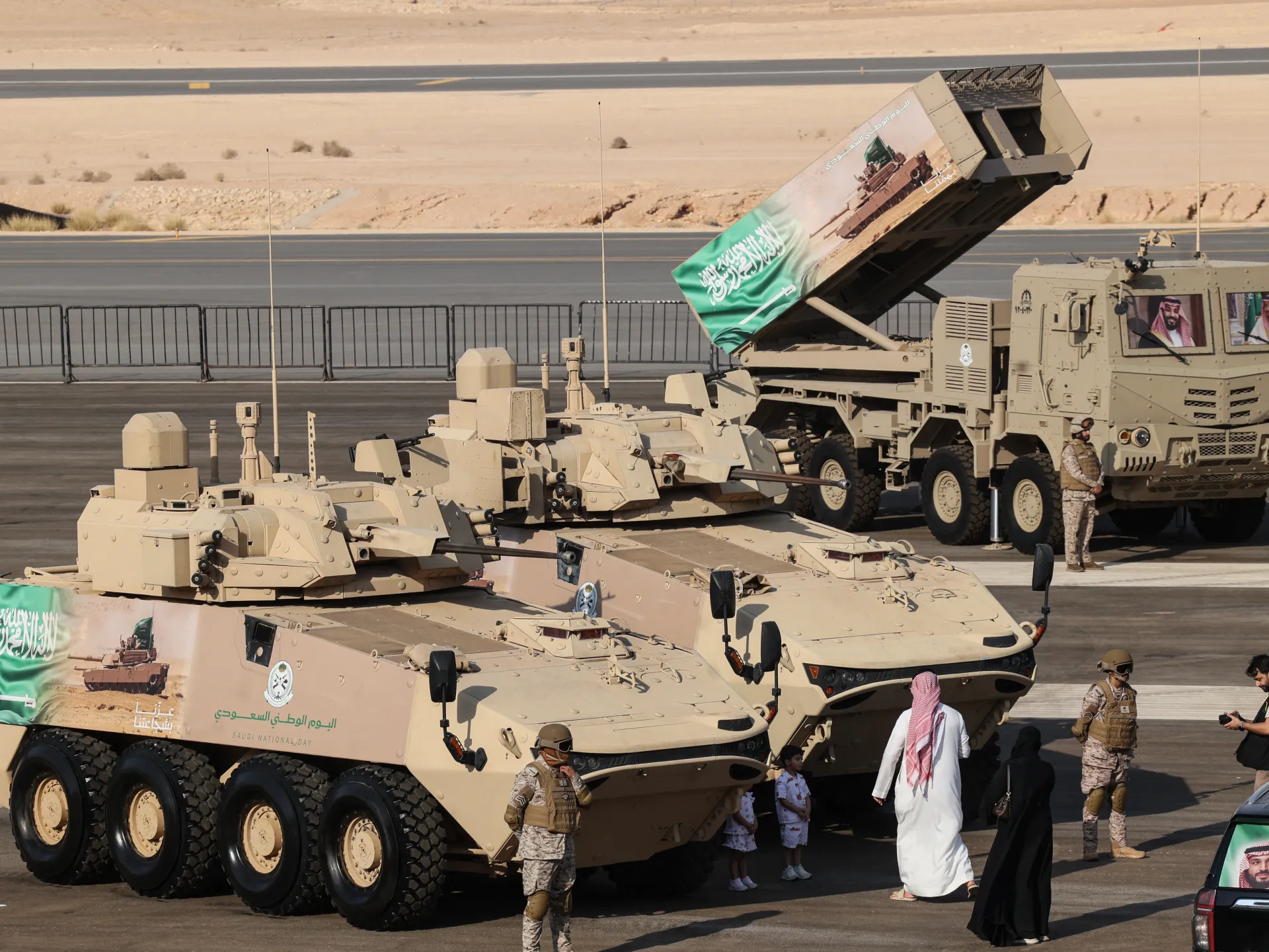 Sand-colored military vehicles and launchers on display as civilians and uniformed personnel stand nearby.