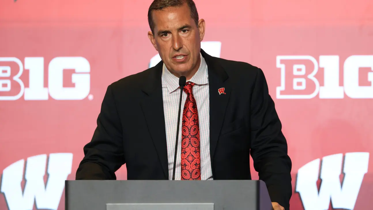 Man in suit and red patterned tie speaking at a podium before a Wisconsin-themed backdrop