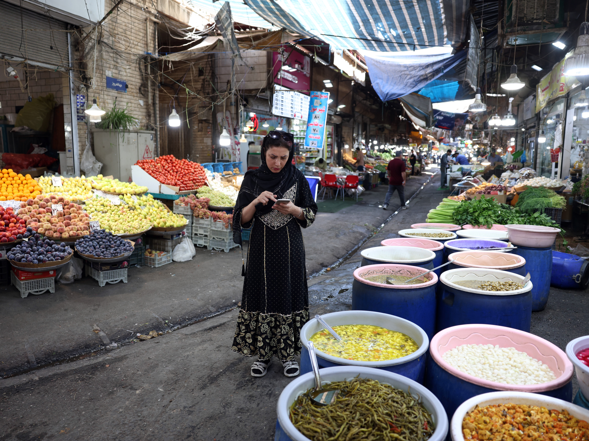 Woman using a smartphone in a covered market surrounded by fruit, vegetables, and barrels of pickled foods.