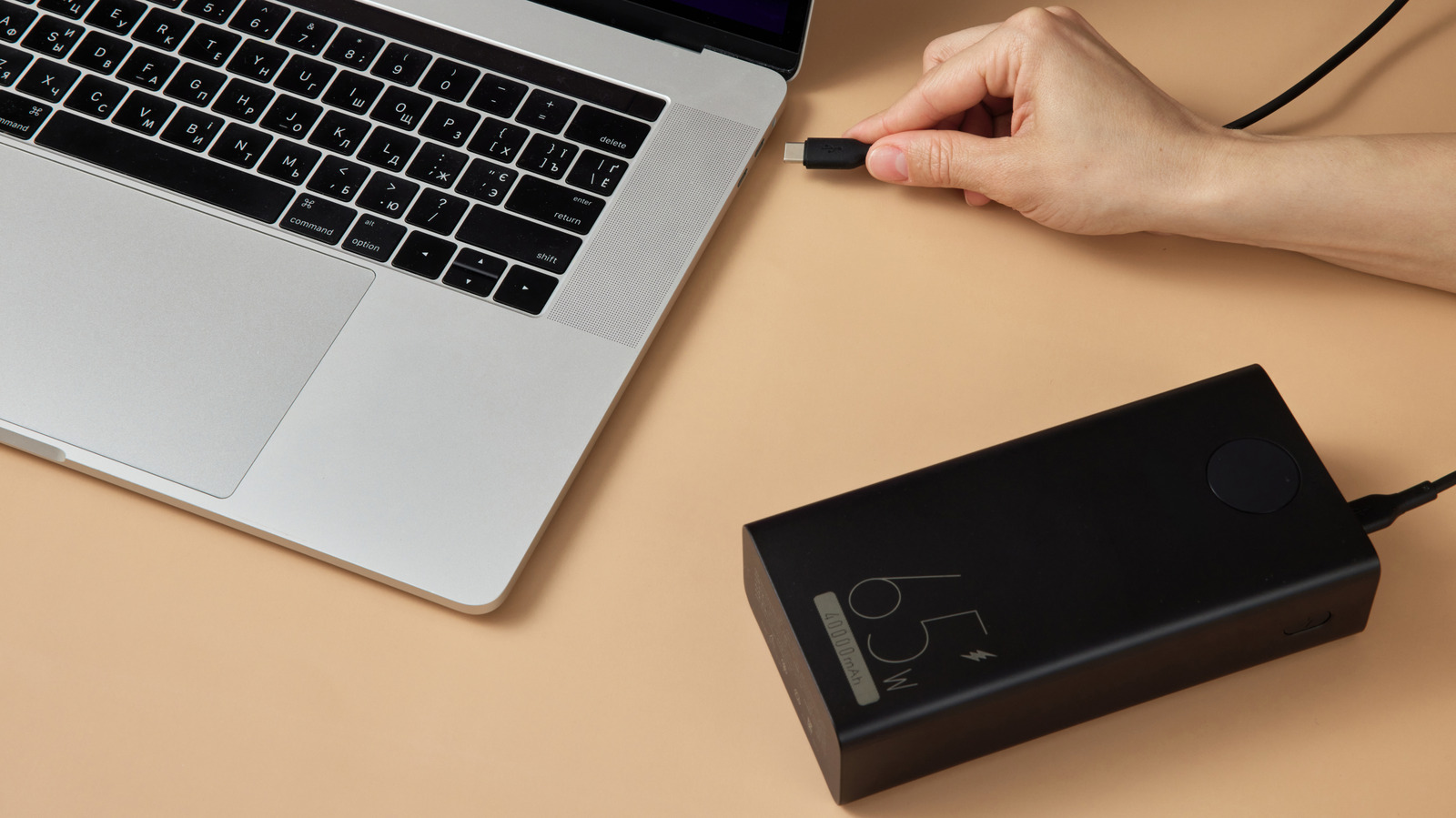 A hand holds a USB-C connector near a silver laptop beside a black power bank on a tan surface.
