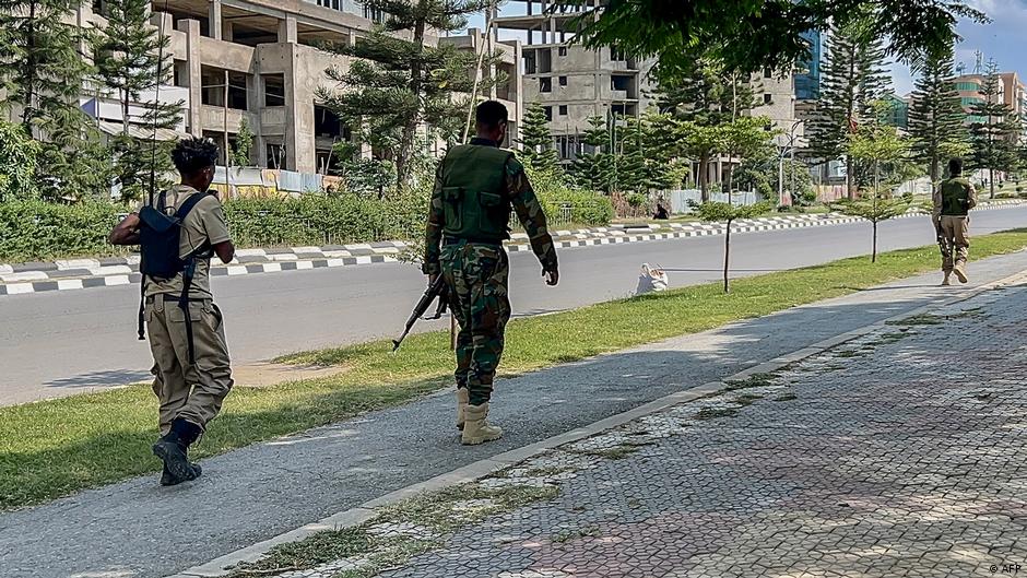 Three armed men walk along a paved path beside a road and modern buildings.