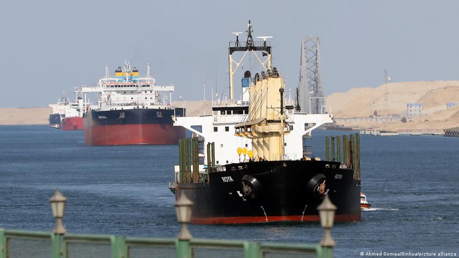 A black cargo vessel sails toward the camera, with larger ships and sandy terrain behind it.