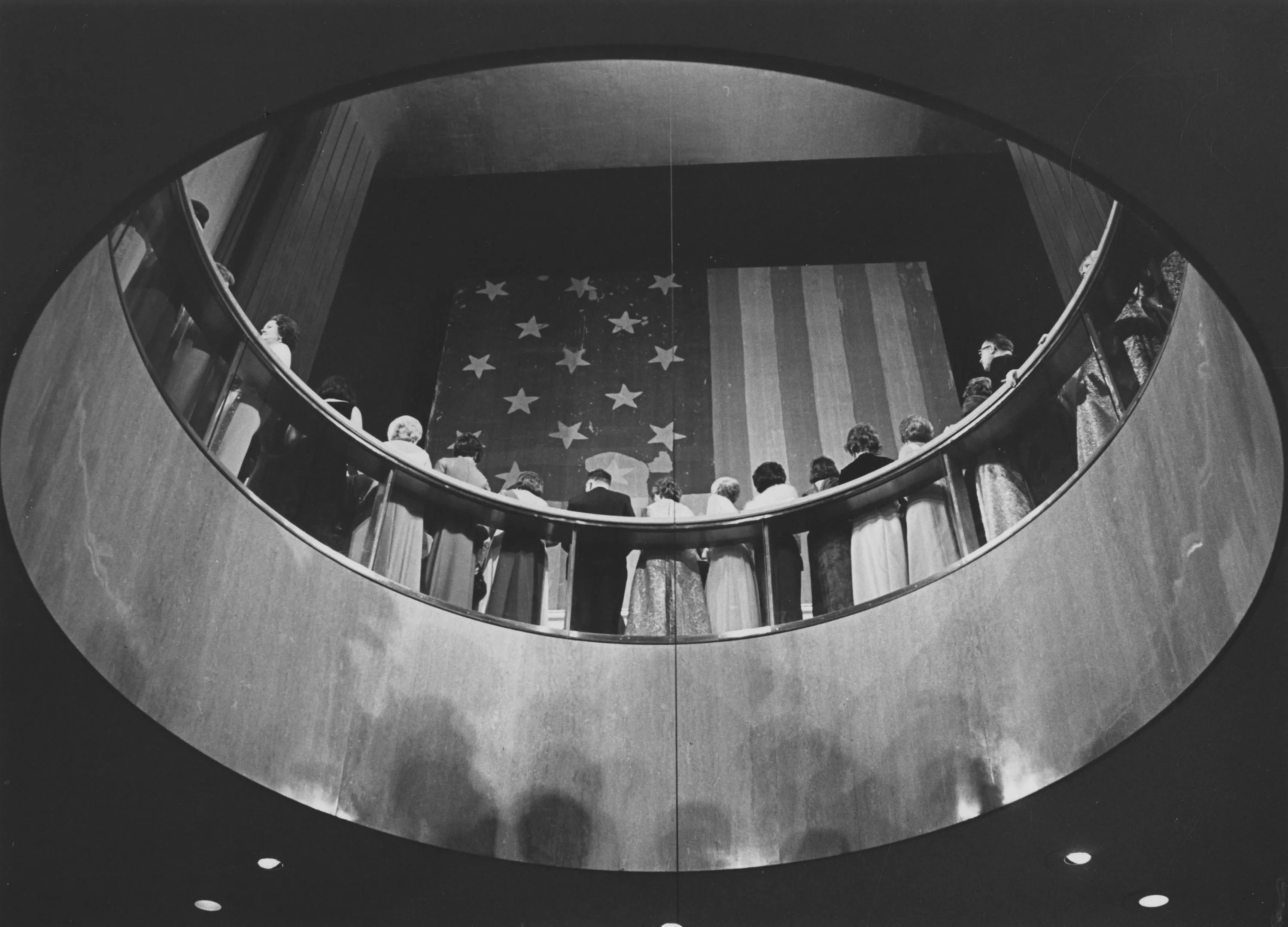 Monochrome view through a curved opening toward people seated before a star-spangled backdrop and American flag.