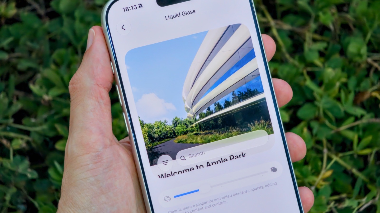 Hand holds an iPhone displaying Liquid Glass settings against a leafy outdoor background.