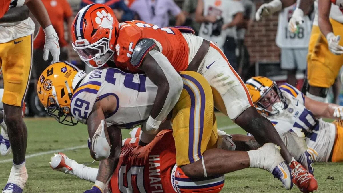 College football players in orange and purple uniforms collide during a tackle on the field.