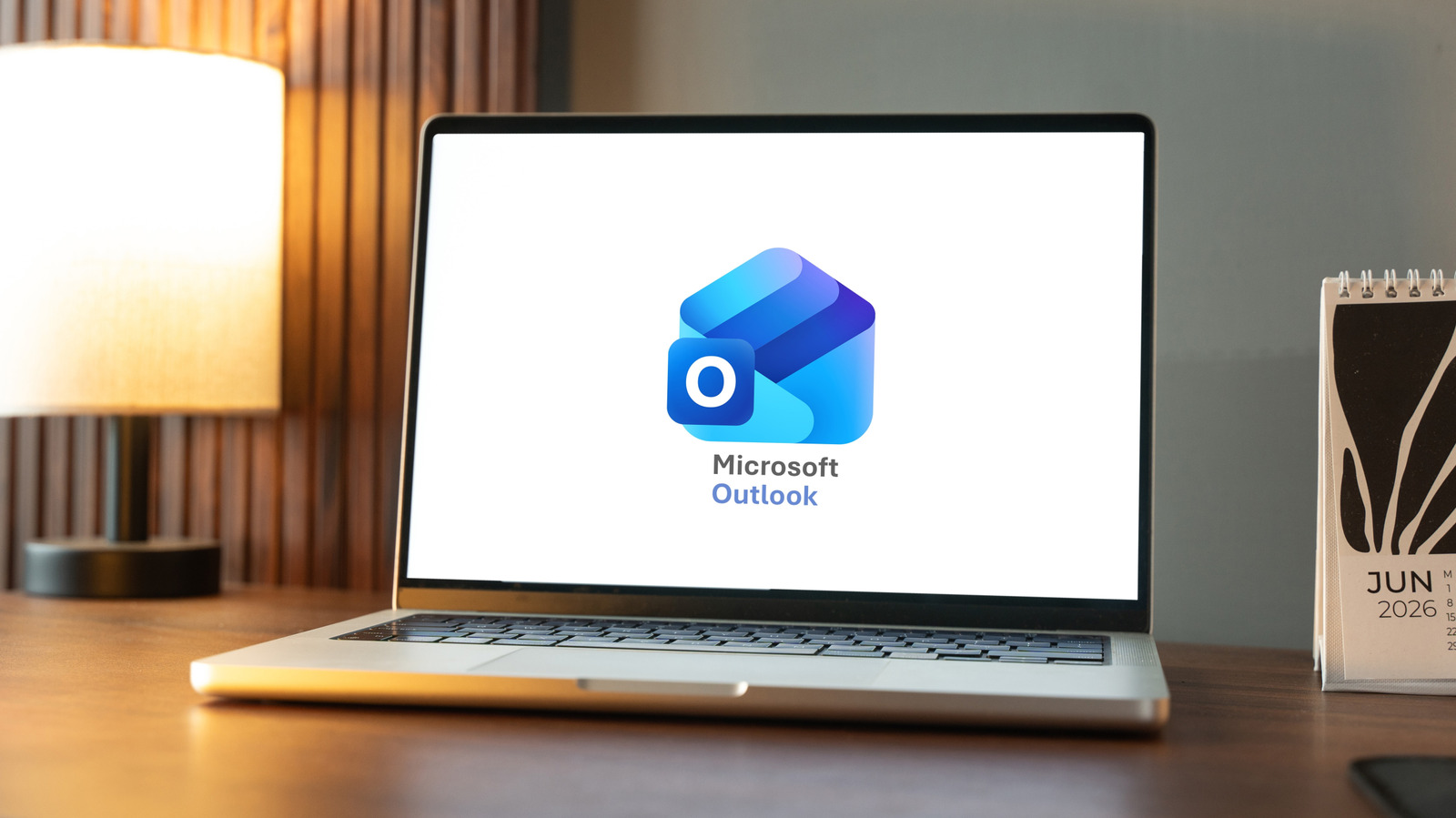 Laptop displaying the Microsoft Outlook logo on a desk beside a lamp and calendar