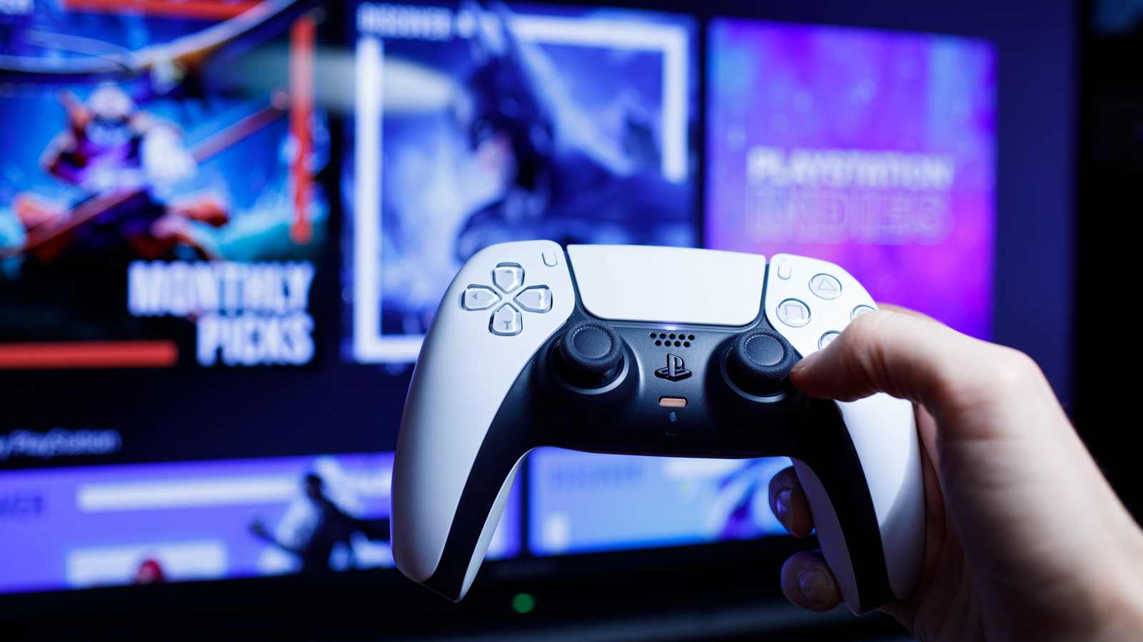 Hand holding a white PlayStation controller in front of colorful video game screens