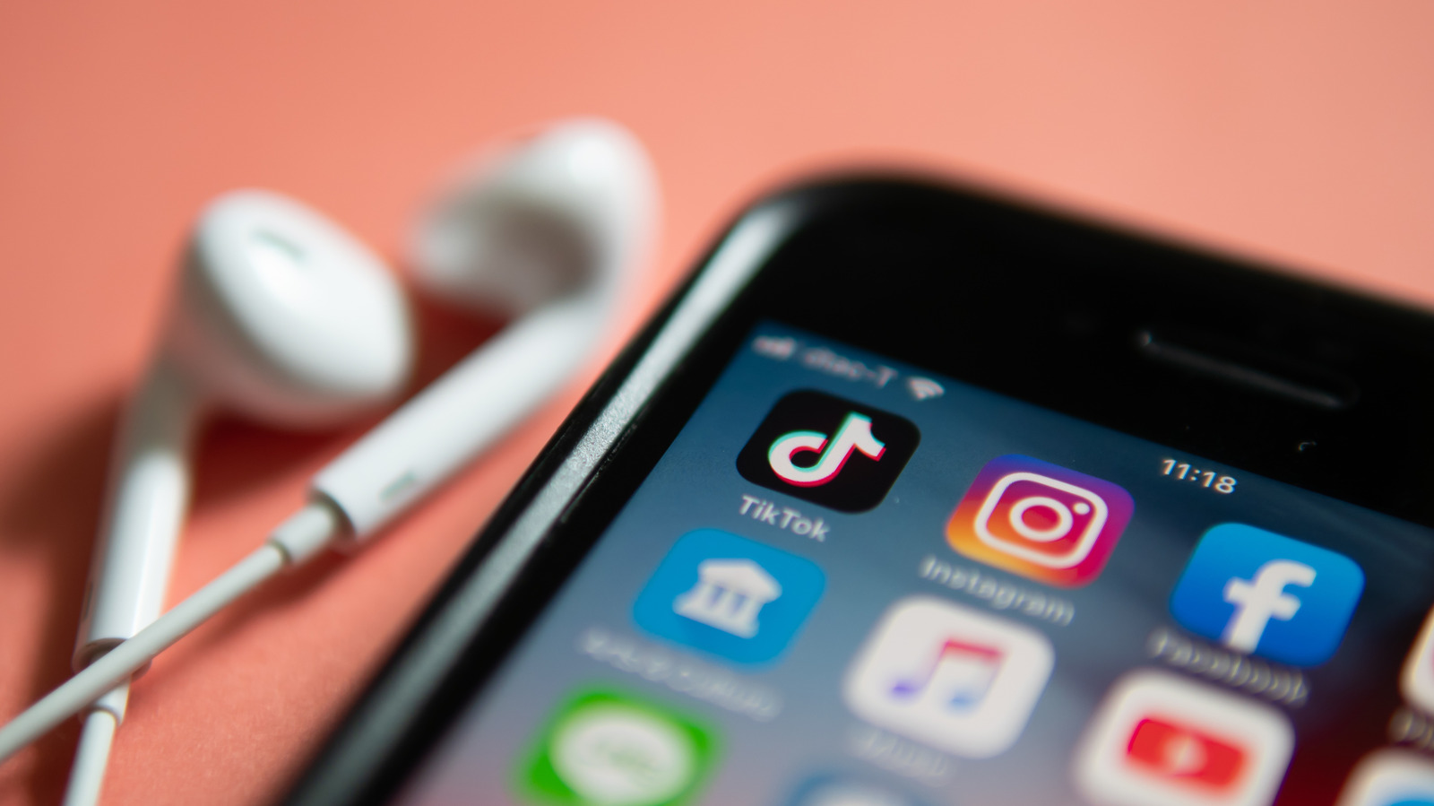 Smartphone displaying social media apps beside white wired earbuds on a coral background