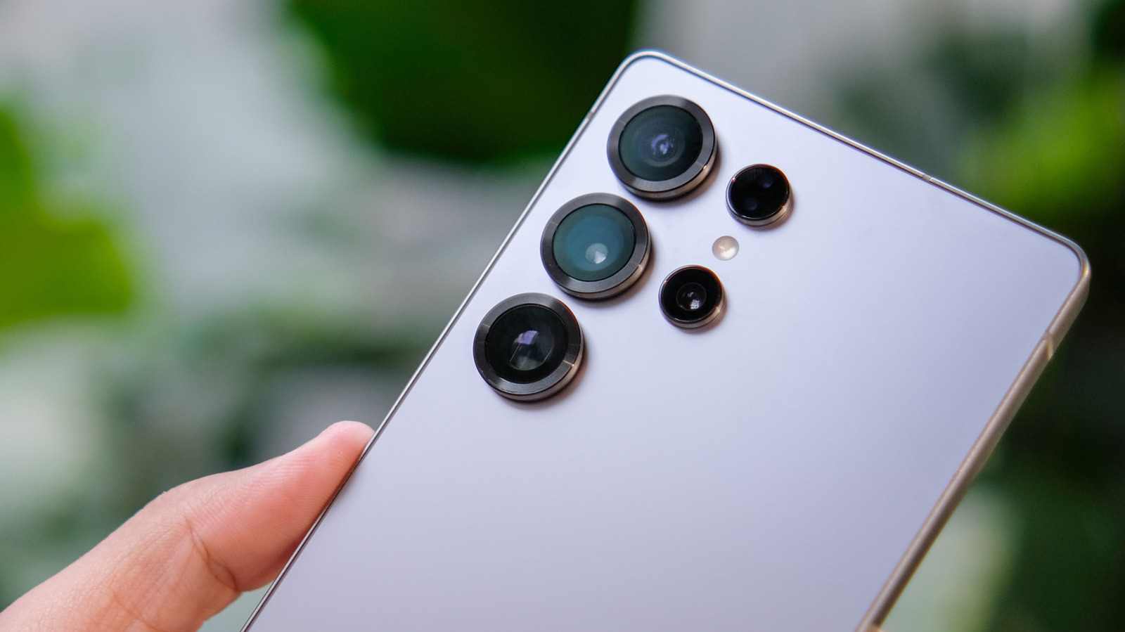 Close-up of a hand holding a pale Samsung smartphone with multiple rear camera lenses against a blurred green background.