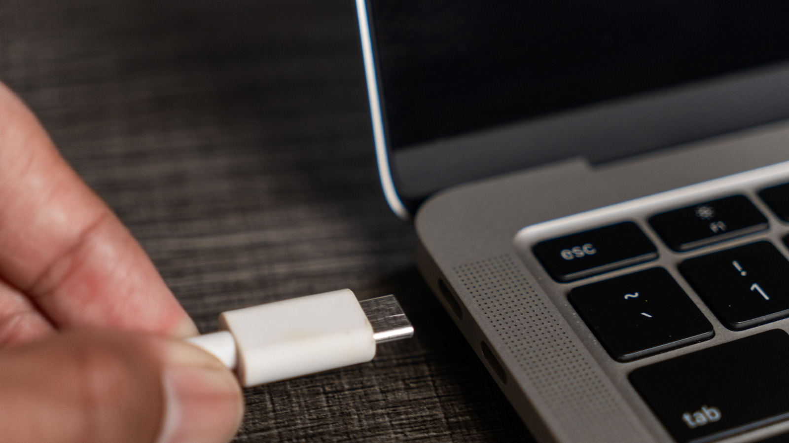 Hand holding a USB-C connector beside a laptop’s USB-C port on a dark desk