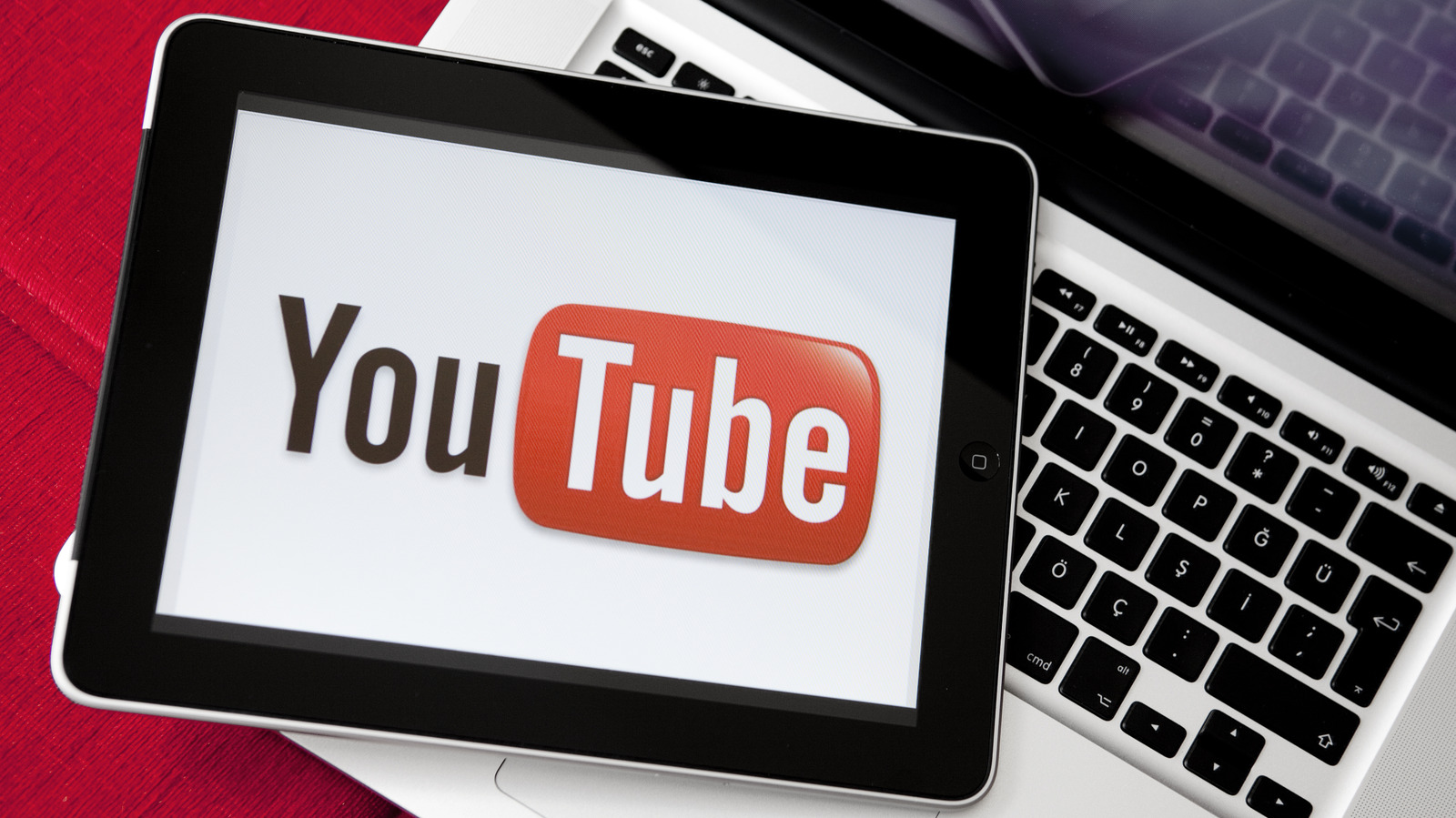 Tablet displaying the YouTube logo resting on a laptop keyboard and red textured surface