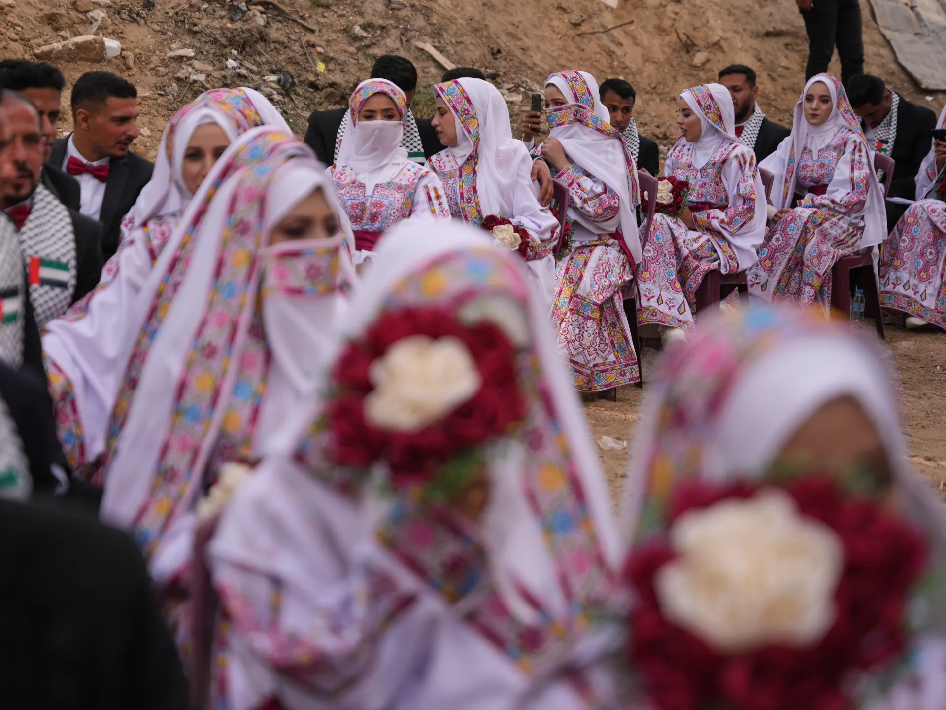 Wedding guests in colorful traditional clothing gather outdoors, with seated women holding bouquets.