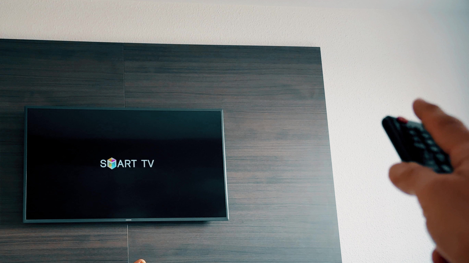 A hand holds a remote control toward a wall-mounted television displaying a Smart TV startup screen.