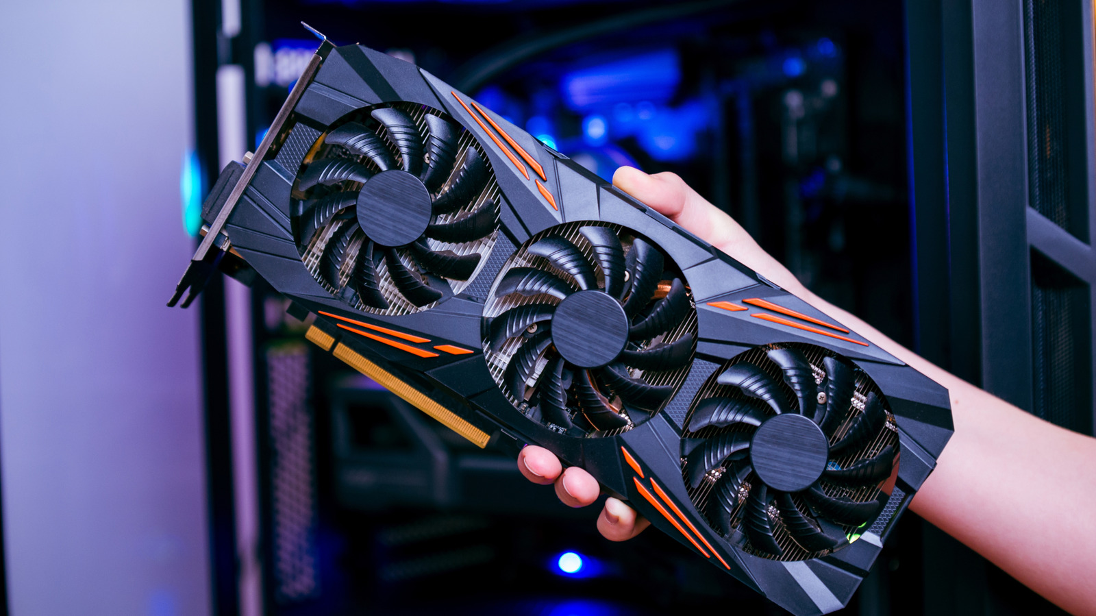 A graphics card with three fans is held in front of a desktop computer.