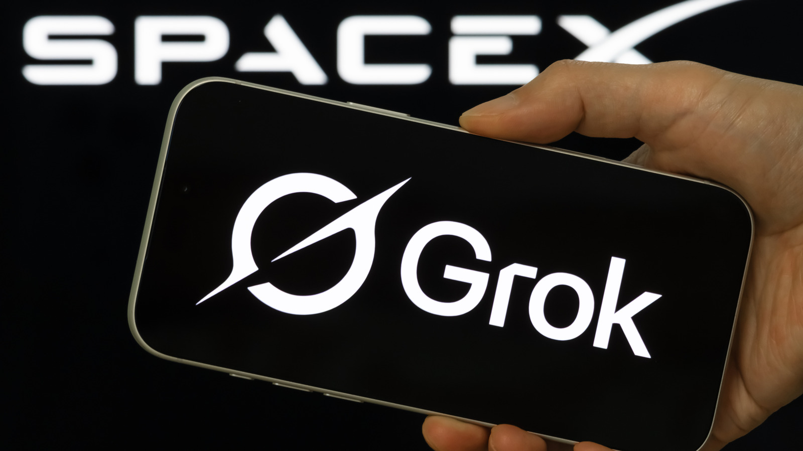 Hand holding a smartphone displaying the Grok logo against a SpaceX sign