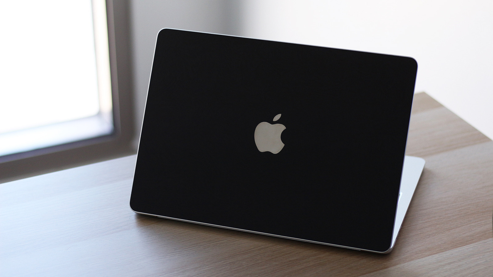 Closed black MacBook displaying the Apple logo on a wooden desk beside a window.