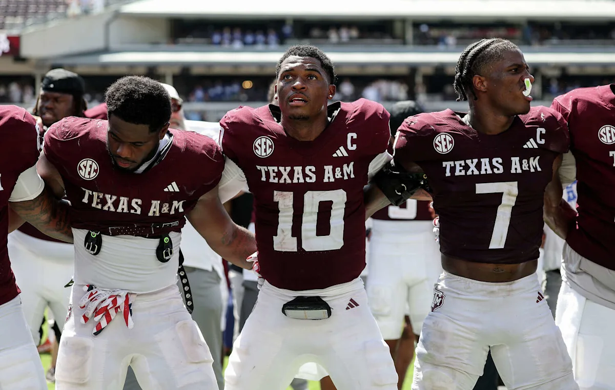 Texas A&M football players stand shoulder to shoulder on the field during a game.