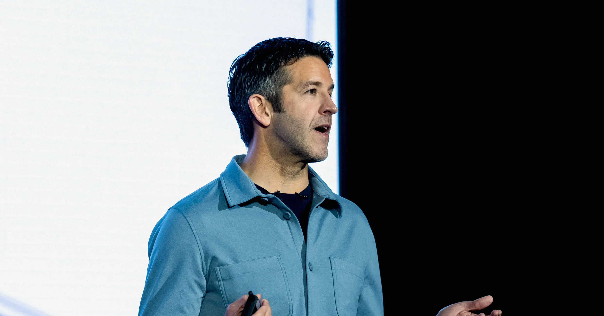 Man in blue jacket speaking onstage while holding a presentation remote