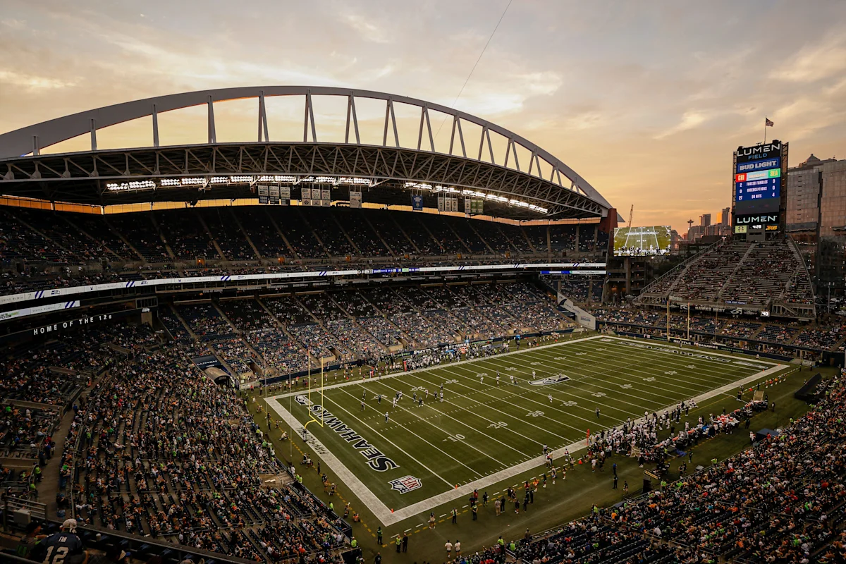 Football stadium filled with fans, field visible beneath arched roof at sunset.