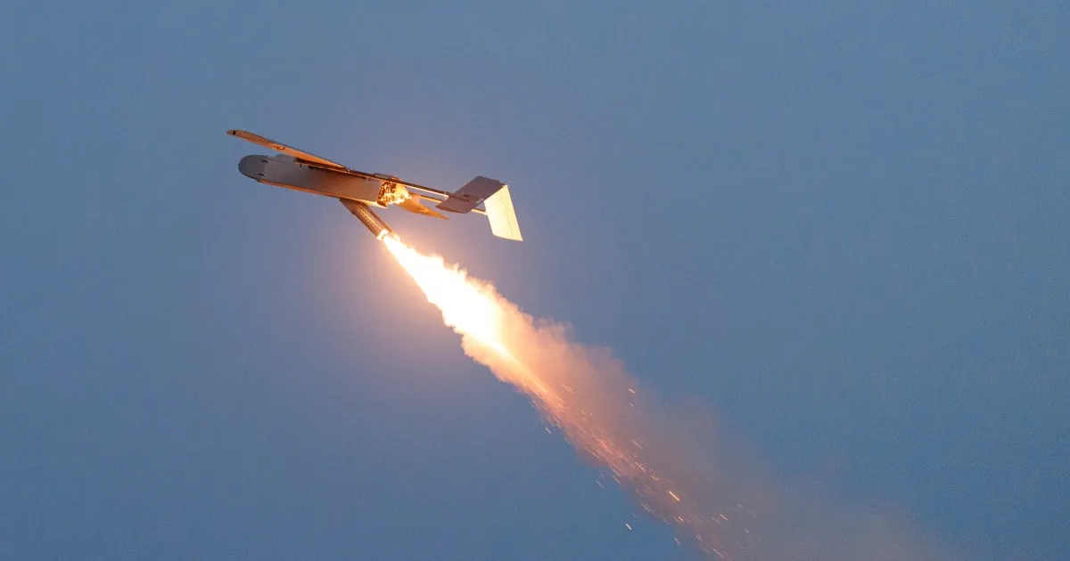 An airborne missile-like object releases a bright flame and smoke trail against a blue sky.