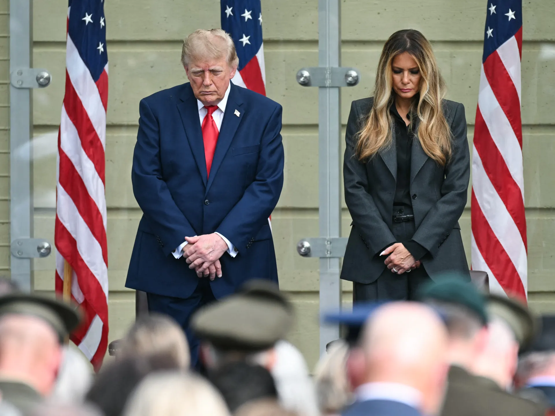 Two formally dressed officials stand solemnly before American flags as a crowd fills the foreground.
