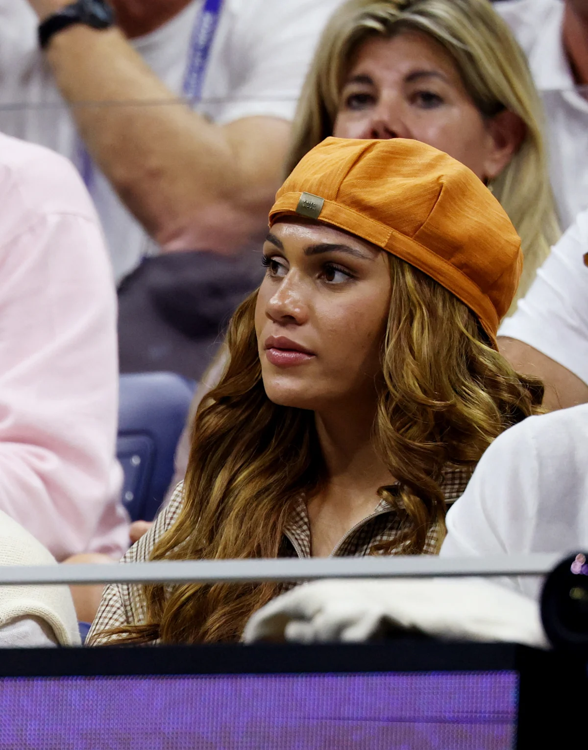 Woman in orange cap watches from stadium seating, with spectators and court barrier around her.