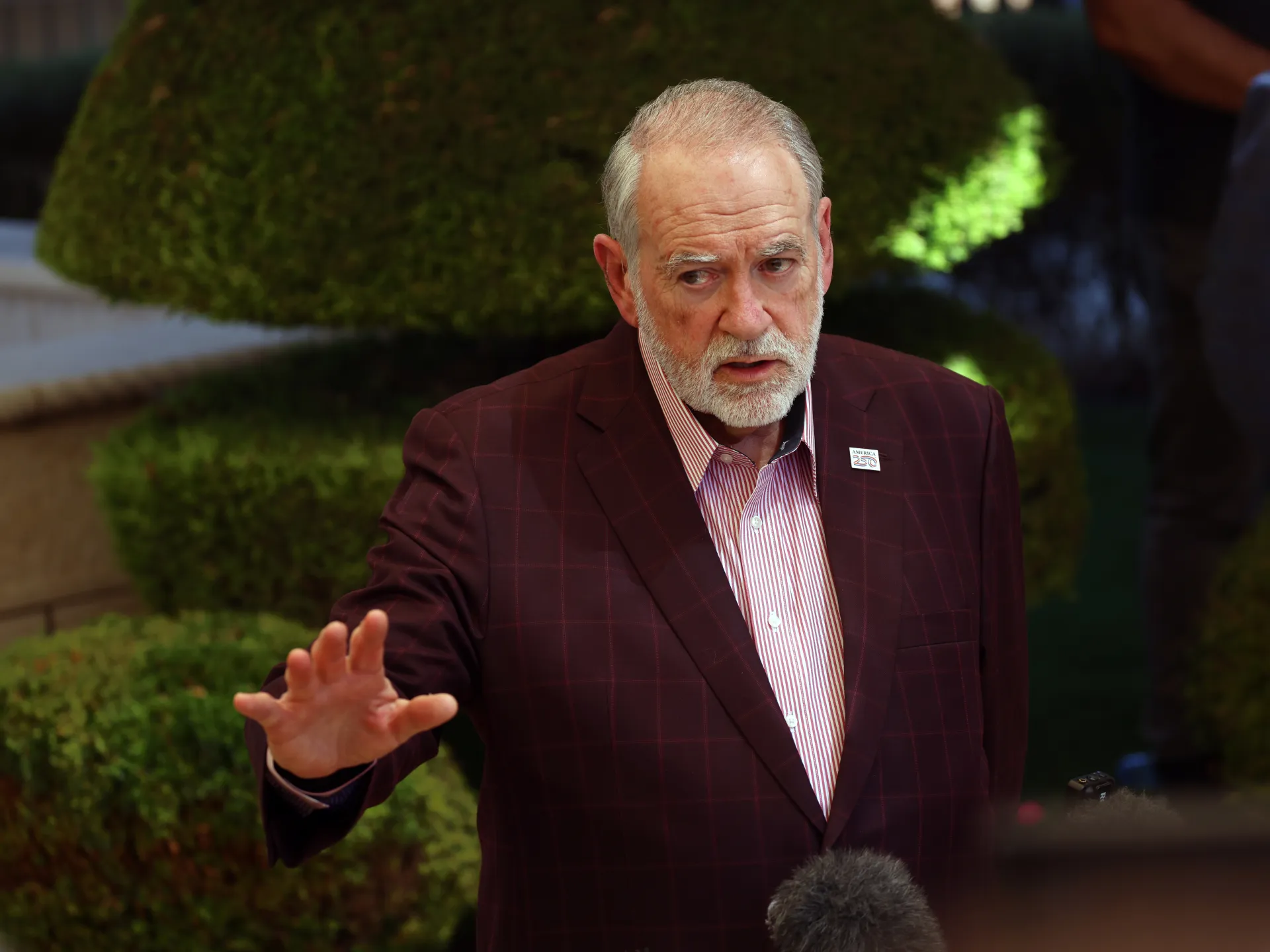 Older man in a burgundy checked blazer gestures toward reporters outdoors, with microphones visible.