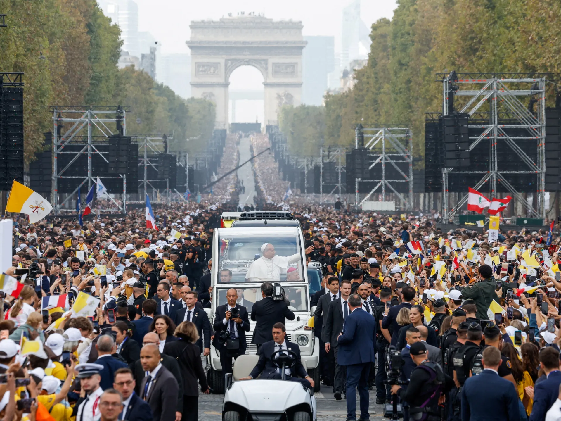 Pope in a vehicle waves to a dense crowd holding flags along a tree-lined avenue.
