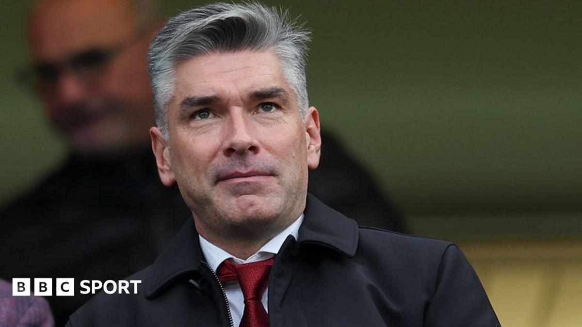 Grey-haired man in a dark coat and red tie looks ahead during a sporting event.